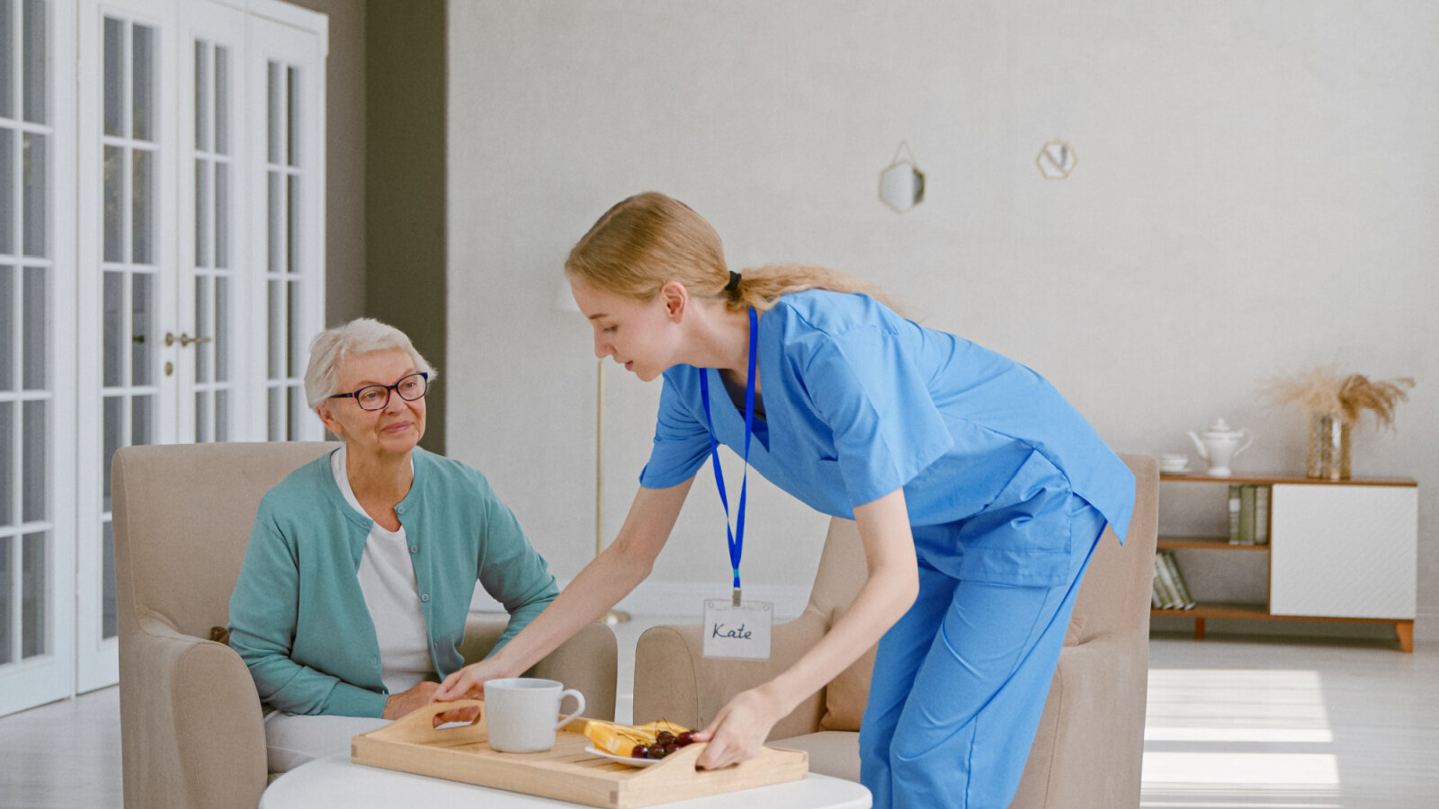 Positive young nurse carries tray with breakfast to senior woman sitting in light living room in Preston Hollow Facilities.