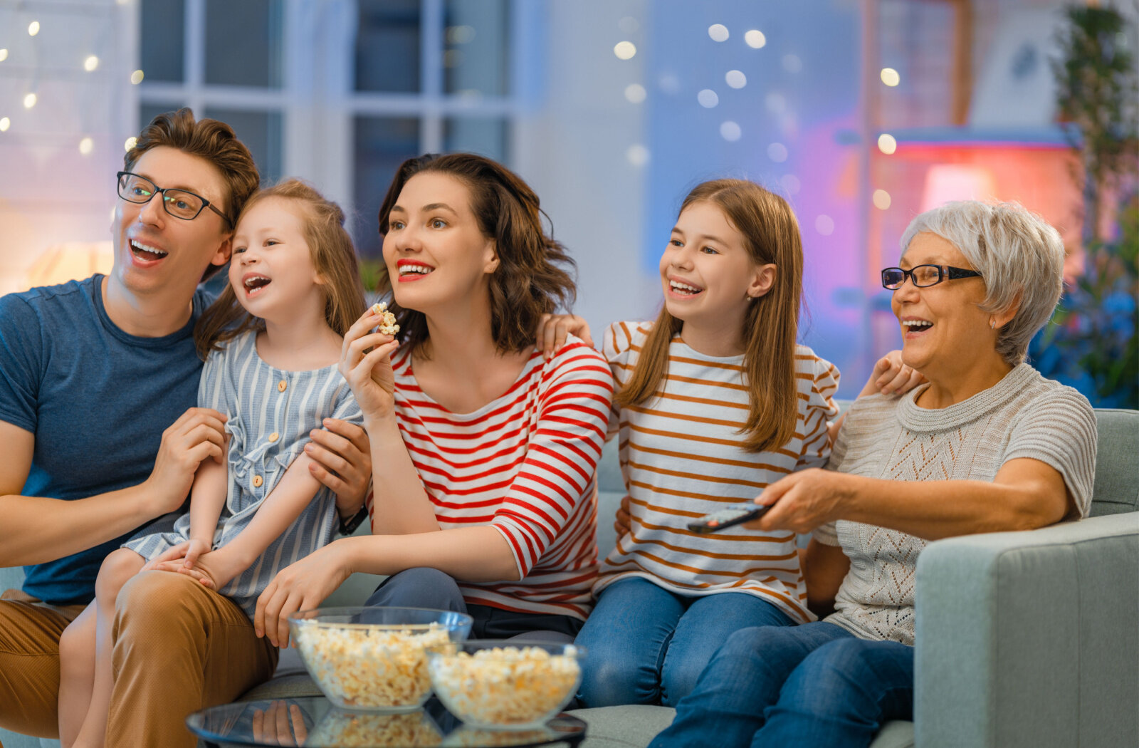 A family sitting on a sofa is spending time together watching movies with the elderly parent.