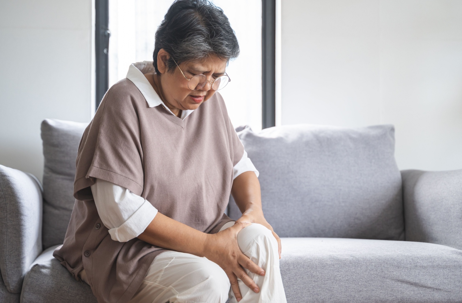 Older woman sitting on a couch holding her knee in pain.