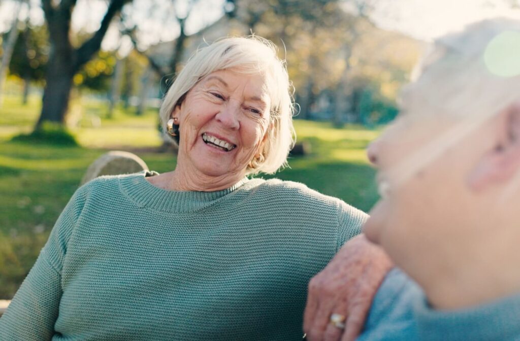 Older adult smiling warmly while chatting with a friend on a bench in a sunny park setting