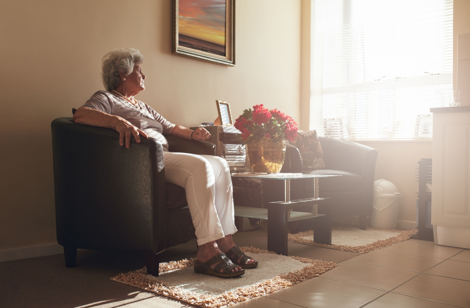 Senior sitting in armchair by window in bright living room with flowers and family photos nearby