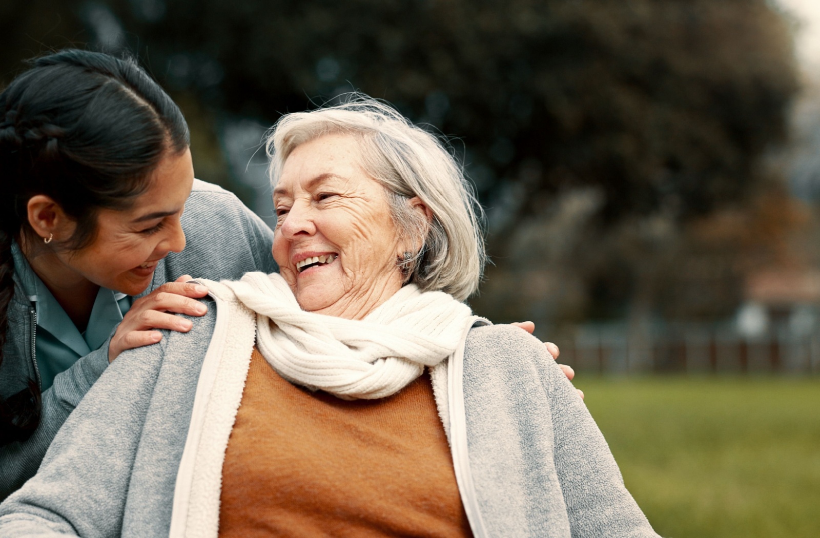 An adult laughs and smiles with their senior parent while outside at their assisted living community during a visit.