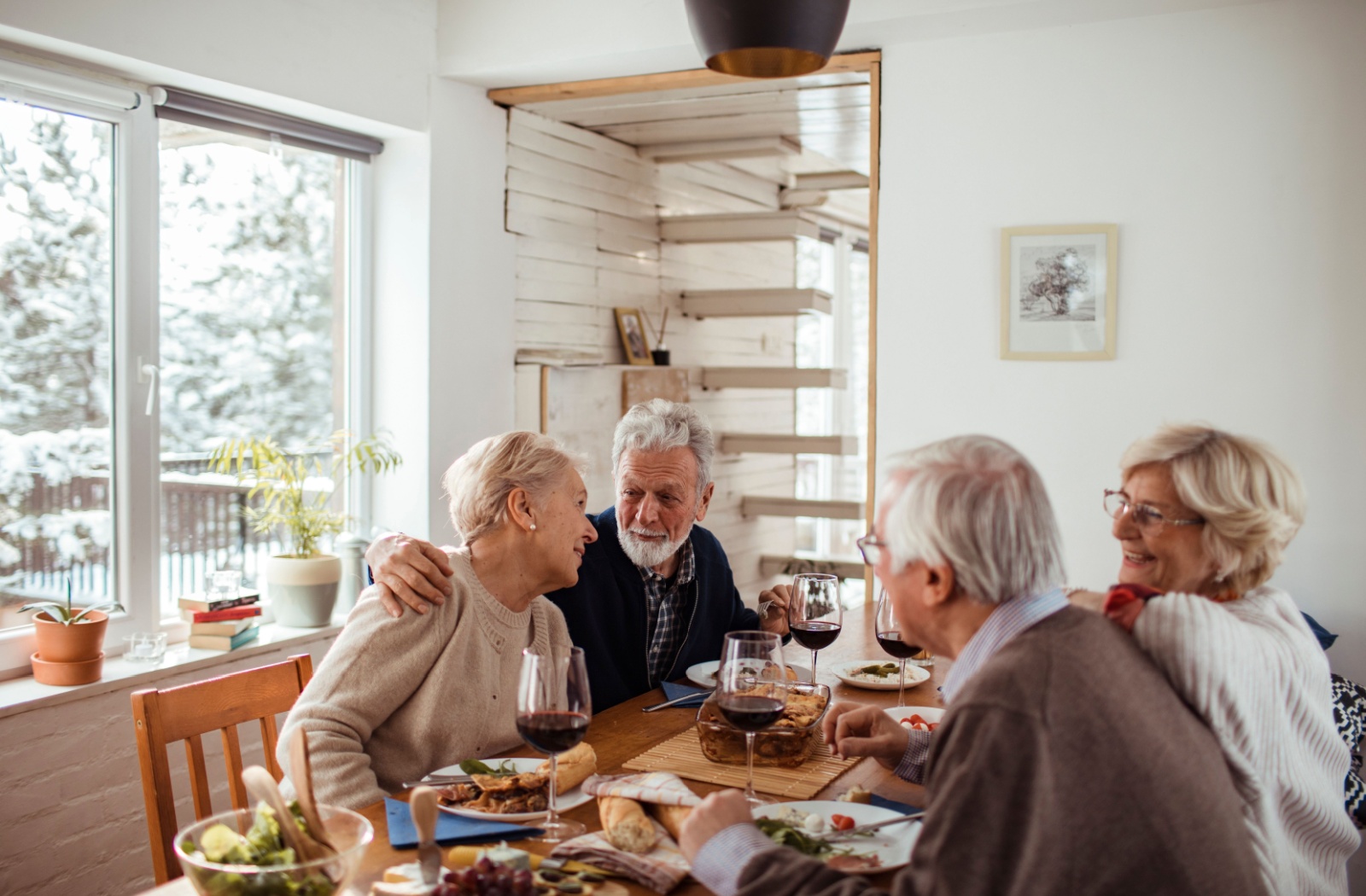 4 older adults laugh over a shared meal in their home in senior living while it snows outside.