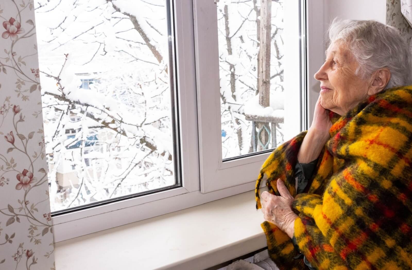 A peaceful older adult wraps themself in a blanket and stares thoughtfully out their window as it snows outside in the winter.